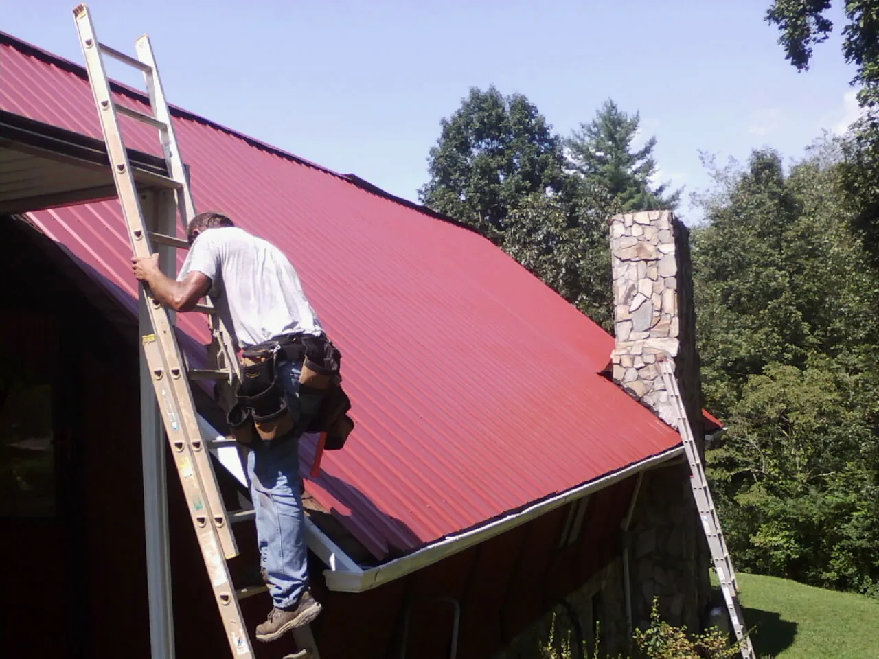 Roofer on a ladder beside a red metal roof and stone chimney — Soffit & Fascia Repair by Roofing Brothers in Donnelsville